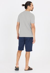 Harmaa lyhythihainen t-paita, tummansiniset shortsit, rento istuvuus. Yksinkertainen muotoilu, jossa on käänteet shortsien helmassa ja ruskeat liukkaat sandaalit. Yksinkertainen tausta.