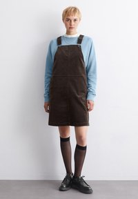 Dunkelbraunes Corduroykleid in A-Linie, verstellbare Träger, kombiniert mit einem hellblauen Sweatshirt und knielangen schwarzen Socken sowie schwarzen Schuhen.