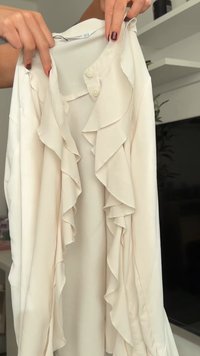 Blusa beige chiaro con dettagli arricciati, chiusura con bottoni e texture liscia. Presenta ampie pieghe di tessuto morbido per una silhouette fluida.