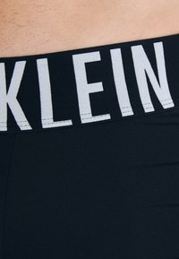 Schwarzer, dehnbarer Stoffbund mit kontrastierendem weißen "KLEIN"-Text. Glatte Textur, schmiegt sich an die Körperlinien an, minimalistisches Design.