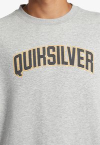 Sweatshirt gris à col rond portant "QUIKSILVER" en lettres noires et orange en gras sur la poitrine. Texture douce.
