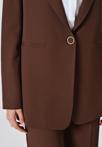 Blazer marrone su misura con bottone dorato arrotondato, linee pulite, tessuto morbido, senza motivi visibili, con dettagli delle tasche laterali e una vestibilità rilassata.
