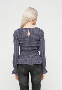 Hosszú szőke hajú nő kék és piros kockás, hosszú ujjú blúzt és fekete nadrágot visel, hátulról látható, egyszínű háttér előtt.