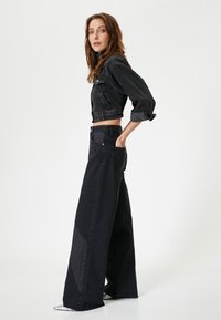 Chaqueta de mezclilla negra corta con cierre de botones y mangas largas, combinada con jeans negros de pierna ancha que presentan detalles de patchwork y bolsillos.