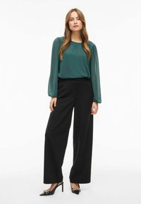Blusa verde de mangas largas con detalle fruncido, combinada con pantalones negros de tiro alto y pierna ancha, y tacones negros de punta con acentos dorados.