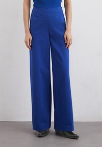 Pantalons larges bleu en tissu lisse, avec une taille haute, des plis sur le devant et une coupe droite. Associés avec des chaussures noires.