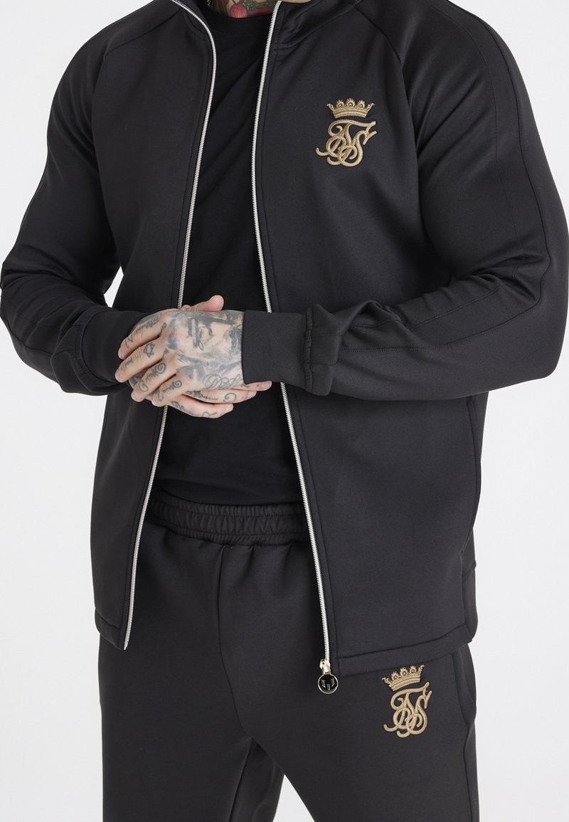 Giacca da corsa nera con logo dorato, chiusura frontale con zip e polsini a coste. Abbinata a pantaloni jogger neri coordinati con simili dettagli dorati.