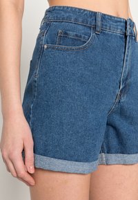 Džínové kraťasy v tmavě modré barvě s vysokým pasem, ohrnutým lemem a designem pěti kapes. Mají bronzové kovové prvky a hladkou texturu.