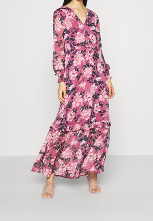 Femme portant une longue robe fleurie rose avec des manches longues transparentes et des détails à volants, associée à des talons à lanières lavande.