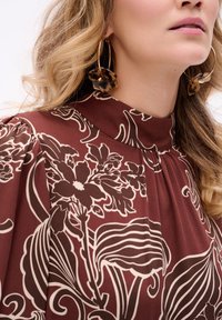 Bordowa bluzka w kwiaty z kremowymi, wirującymi wzorami, z wysokim kołnierzykiem i delikatnymi plisami. Modelka nosi duże okrągłe kolczyki z akcentami w głębi tortoiseshell.