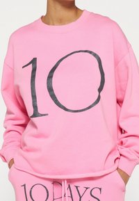 Sudadera de algodón rosa brillante con un gráfico negro de "10". Cuenta con un cuello redondo y puños acanalados, es parte de un conjunto a juego con pantalones jogger.