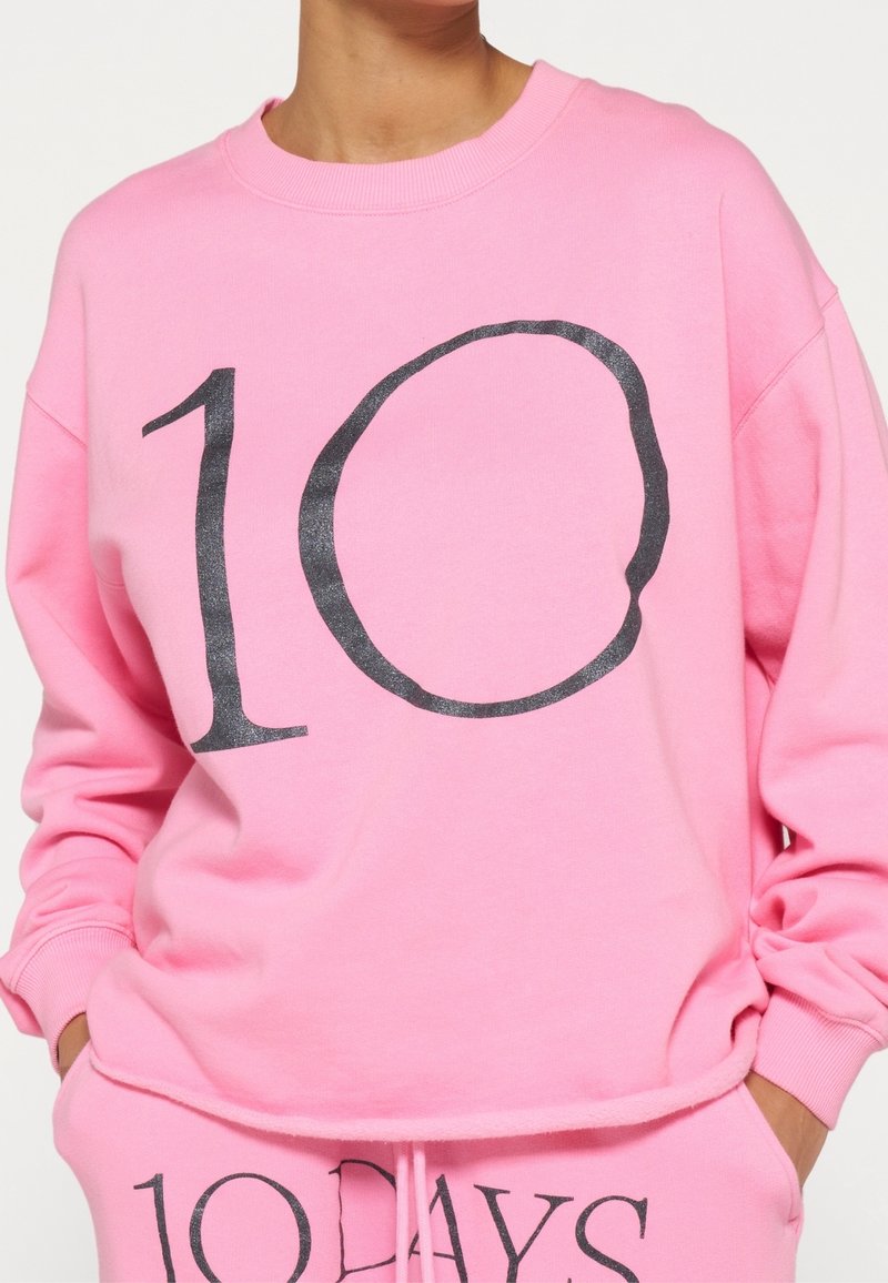 Sudadera de algodón rosa brillante con un gráfico negro de "10". Cuenta con un cuello redondo y puños acanalados, es parte de un conjunto a juego con pantalones jogger.
