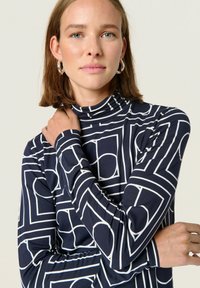 Marineblå langærmet bluse med høj hals, der har et hvidt geometrisk mønster af rektangler og cirkler. Blødt, strækbart stof.