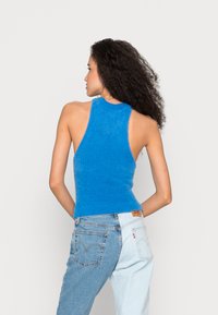 Top de malha azul com um decote alto e costas abertas, apresentando uma textura canelada. Combinado com jeans claros de cintura alta.