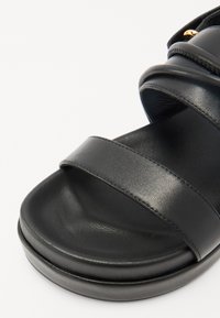 Melnas ādas sandales ar platām siksnām, mīkstu zoli un noapaļotu pamatni. Gluda tekstūra ar redzamām šuvēm.