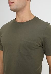 Muž v olivově zeleném tričku s kulatým výstřihem a malou kapsičkou na hrudi, zobrazený od krku po střed trupu na jednobarevném pozadí.