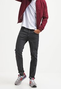 Harmaat slim-fit farkut, joissa on sileä pinta, yhdistettynä burgundinväriseen bomber-takkiin ja harmaisiin lenkkikenkiin, joissa on punaisia ja sinisiä yksityiskohtia.