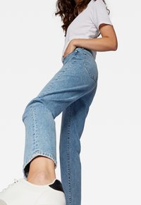 Vaqueros de mezclilla azul claro con cintura alta, diseño de pierna recta y desvanecimiento sutil. Combinados con zapatillas blancas con un detalle en negro.