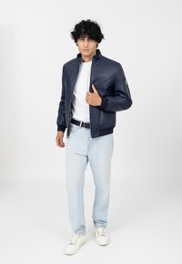 Veste bombardier en cuir bleu marine avec des poignets et un col côtelés, poche zippée sur la manche, portée par-dessus un T-shirt blanc et un jean bleu clair.