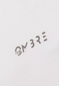 Bílý tričko s šedým tištěným logem a textem "OMBRE LEVEL UP", použitím moderního, geometrického písma. Hladká bavlněná textura.