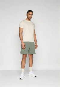 Camiseta beige, pantalones cortos verdes, zapatillas blancas y calcetines crema hasta el tobillo. El conjunto presenta un diseño casual con color liso y ajuste relajado.