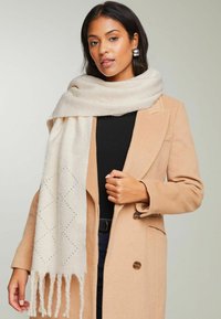 Beige sjaal van zachte stof, met diamantpatronen en franjes aan de uiteinden, gedragen over een zwart shirt en een camelkleurige jas. Elegante uitstraling.