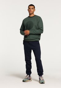 Groene sweatshirt met een gestructureerd cirkelontwerp, gecombineerd met marineblauwe joggers. Het model draagt kleurrijke sneakers, met ontspannen armen langs de zij.