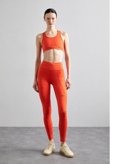 Bright orange activewear set, featuring a cropped sports bra and high-waisted leggings, with a smooth texture and logo accents. Finished with beige sneakers.