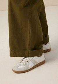 Des baskets blanches et beiges avec une semelle en gomme, fabriquées en daim et en cuir. Associées à un pantalon large vert olive.