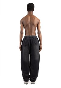 Črne hlače ohlapnega kroja z teksturirano tkanino, elastičnim pasom in stranskimi žepi, nošene z sivimi športnimi čevlji. Pogled od zadaj.