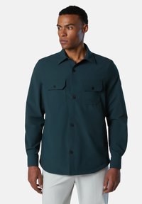 Camisa de manga larga en verde azulado oscuro con cuello abotonado, dos bolsillos en el pecho y un corte relajado. Tejido suave con un ligero brillo.