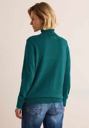 Turkos polotröja i mjukt stickat material, med lång ärm och ribbade ärmslut. Bärs med blå denimjeans, vy från baksidan.
