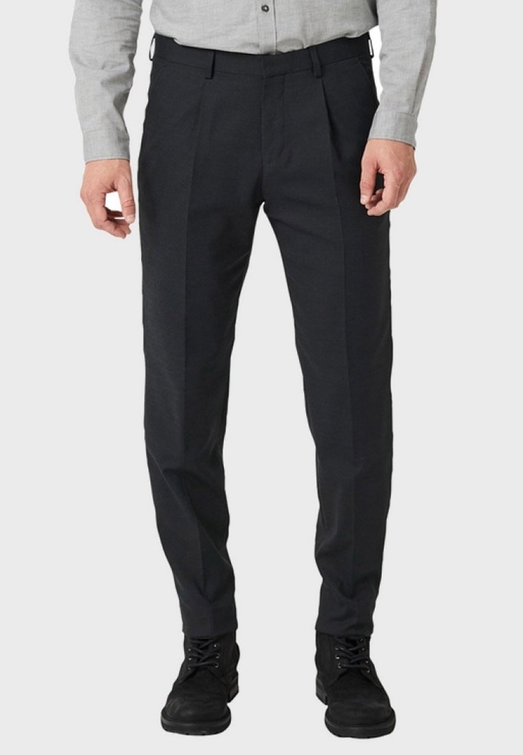 Homme portant un pantalon de costume noir, une chemise gris clair boutonnée et des chaussures noires à lacets sur un fond clair uni.