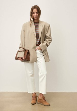 Naine kannab üle suuruses beeži bleiserit, triibulist pruuni toppi, valgeid pükse, beeže spordijalatsid, hoiab pruuni käekotti, seisab ühevärvilise tausta ees.