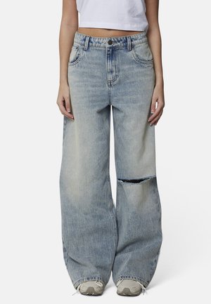 Donna che indossa jeans azzurri a gamba larga e strappati con una singola rottura al ginocchio, top bianco corto e sneakers bianche su sfondo semplice.