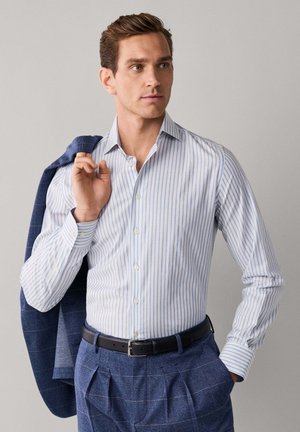 Homme en chemise rayée bleu et blanc, pantalon à carreaux bleus et veste posée sur l’épaule, regardant vers la droite.
