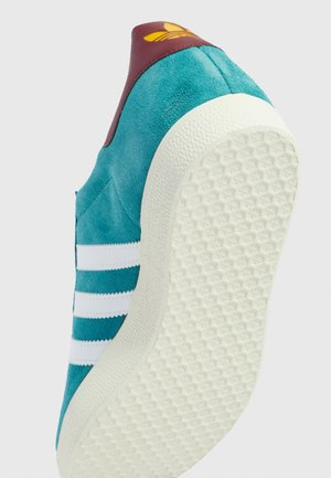 Tyrkisblå ruskindssneakers med hvide striber, tekstureret hvid sål og bordeaux hælflap med gult trebladet logo.