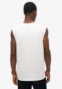 Camiseta sin mangas de algodón blanco con un escote redondeado y una textura suave, con costuras visibles en el dobladillo y un logo sutil en la parte posterior.