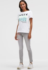 Weißes Baumwoll-T-Shirt mit buntem Grafiktext, kombiniert mit hellgrauen Slim-Fit-Jeans mit aufgedruckten Akzenten. Die Schuhe sind voluminöse weiße Sneaker.