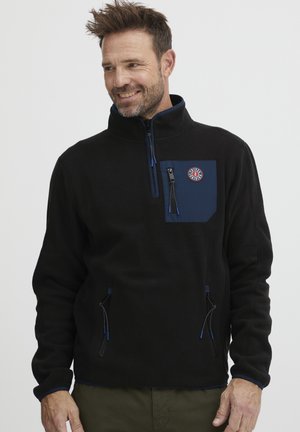 Černý flísový pulovr s vysokým límcem, modrou kapsou na hrudi, dvěma kapsami na zip po stranách a kontrastními modrými detaily na zipsech.