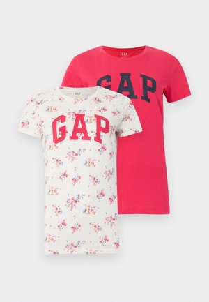 Žena u bijeloj majici s cvjetnim uzorkom i natpisom "GAP", svijetloplavim širokim trapericama s kratkim okovratnicima i crnim baletnim cipelama stoji ispred bijele pozadine.