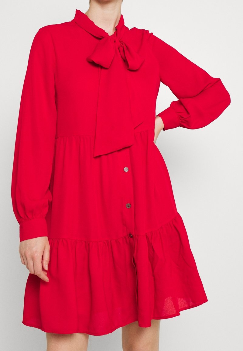 Femme portant une robe rouge à manches longues avec des boutons sur le devant et un grand nœud au niveau de l'encolure.