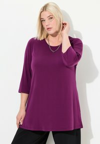 Donna bionda che indossa una tunica a maniche lunghe viola e pantaloni neri, toccandosi i capelli con una mano su uno sfondo bianco semplice.