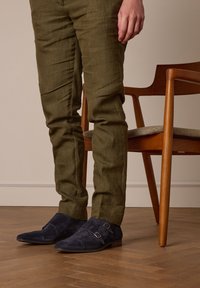 Marineblauwe suède dubbele monk-strap schoenen met een puntige neus, gecombineerd met olijfgroene gestructureerde broeken, gezien tegen een houten vloer en stoel.