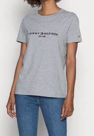 Personne portant un t-shirt gris clair Tommy Hilfiger avec logo et texte "Est. 1985", associé à un jean bleu sur un fond uni.