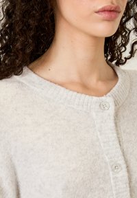 Světle šedý cardigan vyrobený z měkké, texturované látky. Má žebrovaný límec a dva knoflíky s hladkým, zaobleným designem.