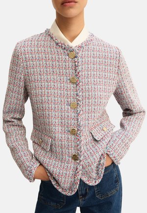 Donna che indossa una giacca di tweed multicolore con bottoni dorati, tasche frontali e scollo rotondo, in piedi con le mani nelle tasche.