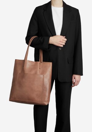 Borsa a mano in pelle marrone con manici lunghi, dotata di una grande tasca frontale e dettagli di cucitura minimal, portata da una persona in un completo nero.