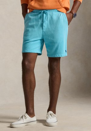 Mężczyzna ubrany w turkusowe szorty ze sznurkiem, białe tenisówki i pomarańczową bluzę z kapturem, z rękami w kieszeniach, stojący na neutralnym tle.