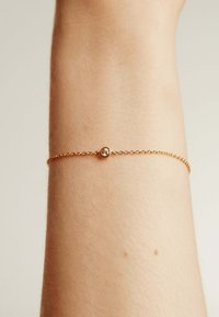 Pulsera de cadena de oro con una pequeña gema clara, descansando delicadamente sobre un tono de piel neutro. Diseño minimalista con una textura fina.